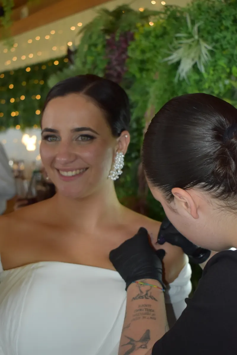 Alba tatuando un mini tattoo en el hombro de una novia sonriente, en una boda con jardín vertical y guirnaldas de luces