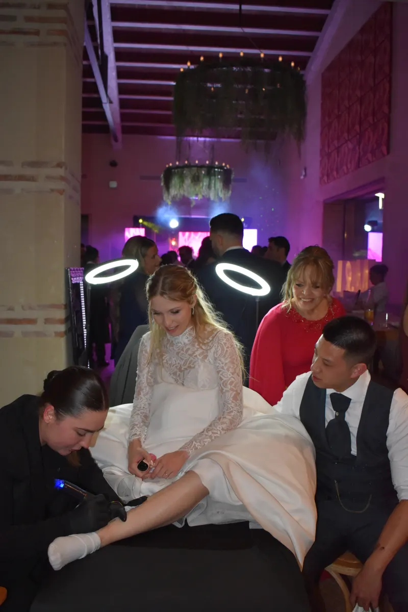 Alba tatuando el tobillo de una novia durante la fiesta de la boda, con el novio y una invitada mirando bajo luces de aro