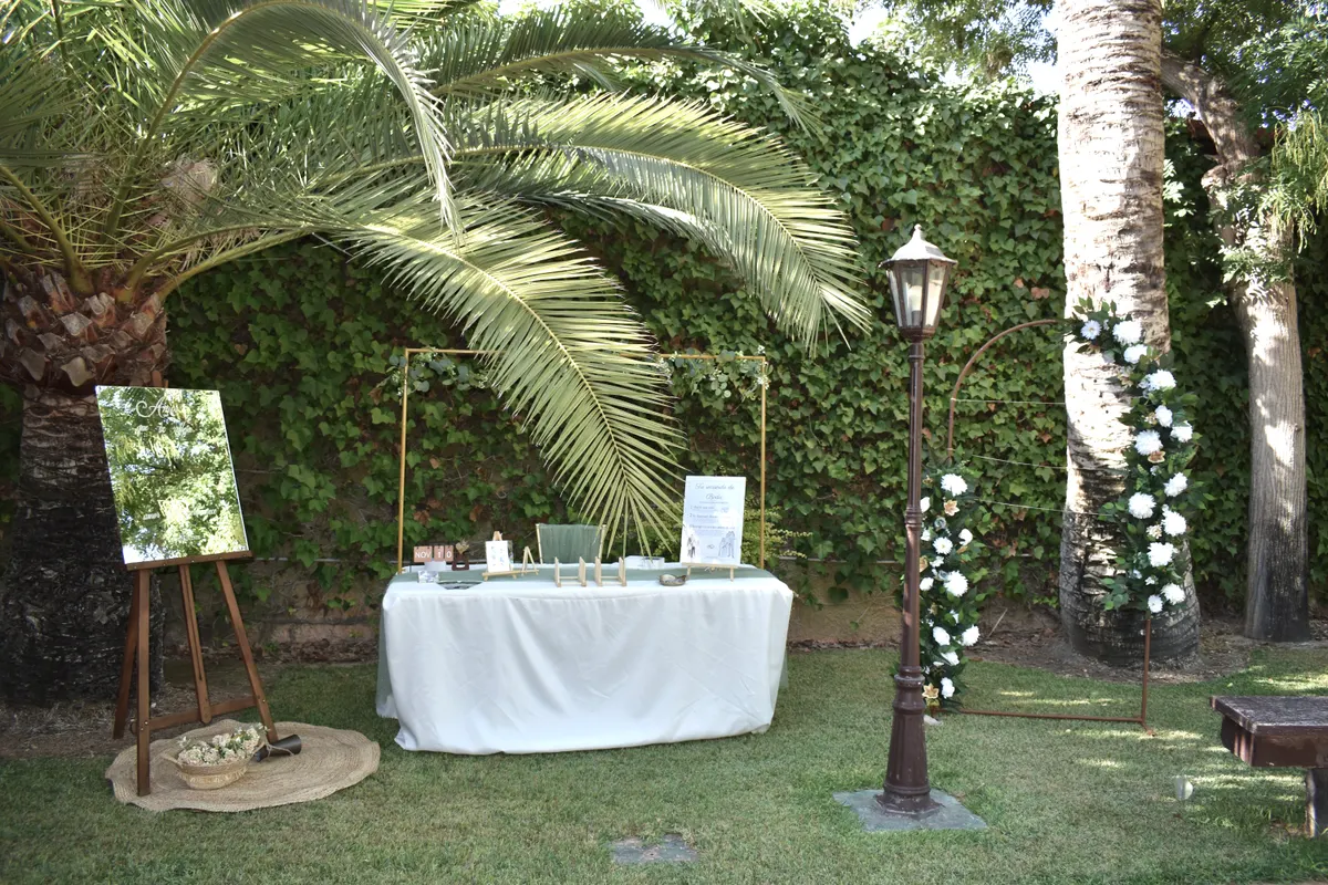 Mesa de ilustración en directo preparada bajo una palmera: mantel blanco, caballete con espejo, marco dorado y arco de flores blancas