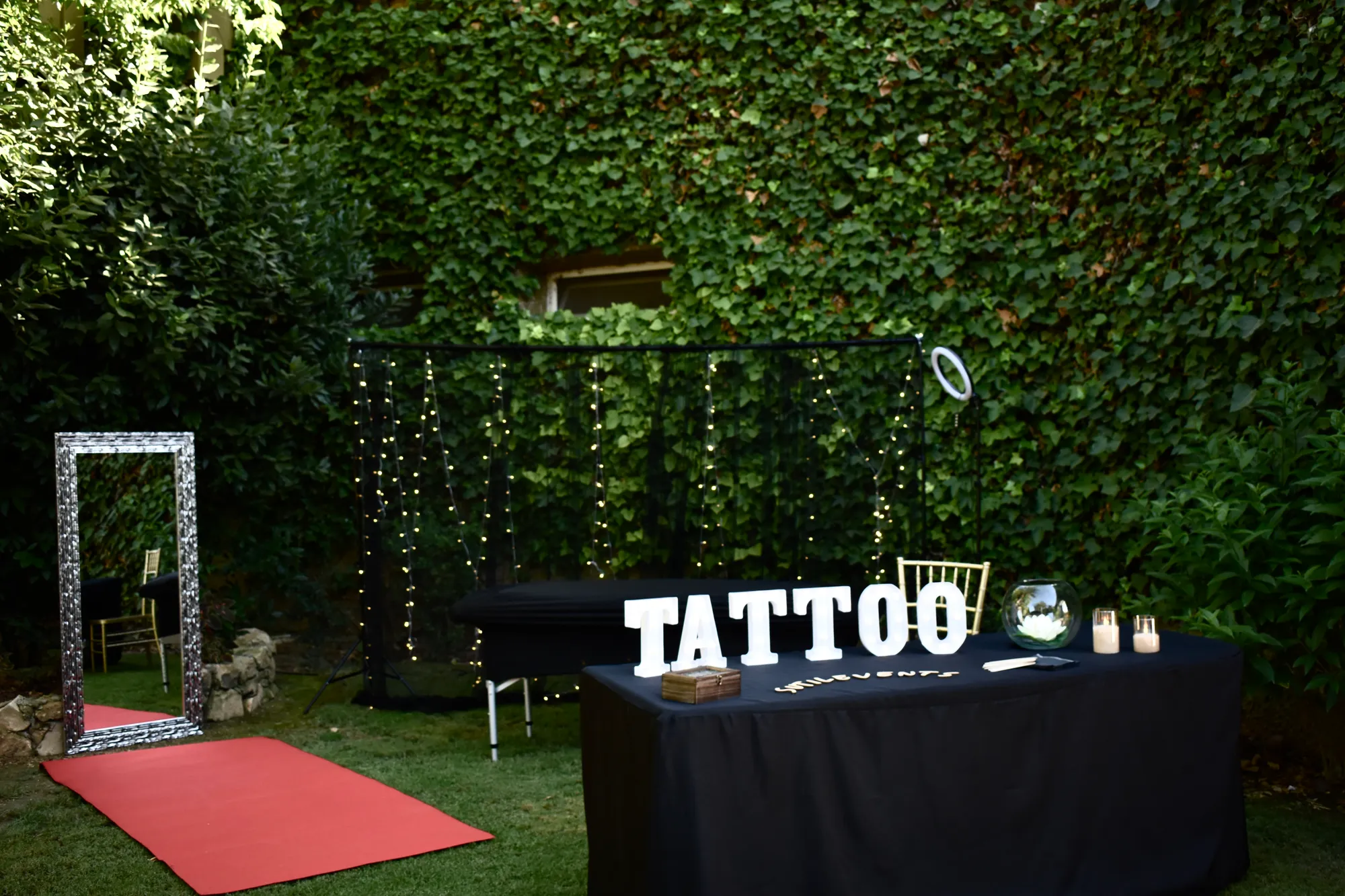 Mini tattoo corner set up in a wedding garden: ivy wall, curtain lights, black couch, TATTOO letters, mirror and red carpet