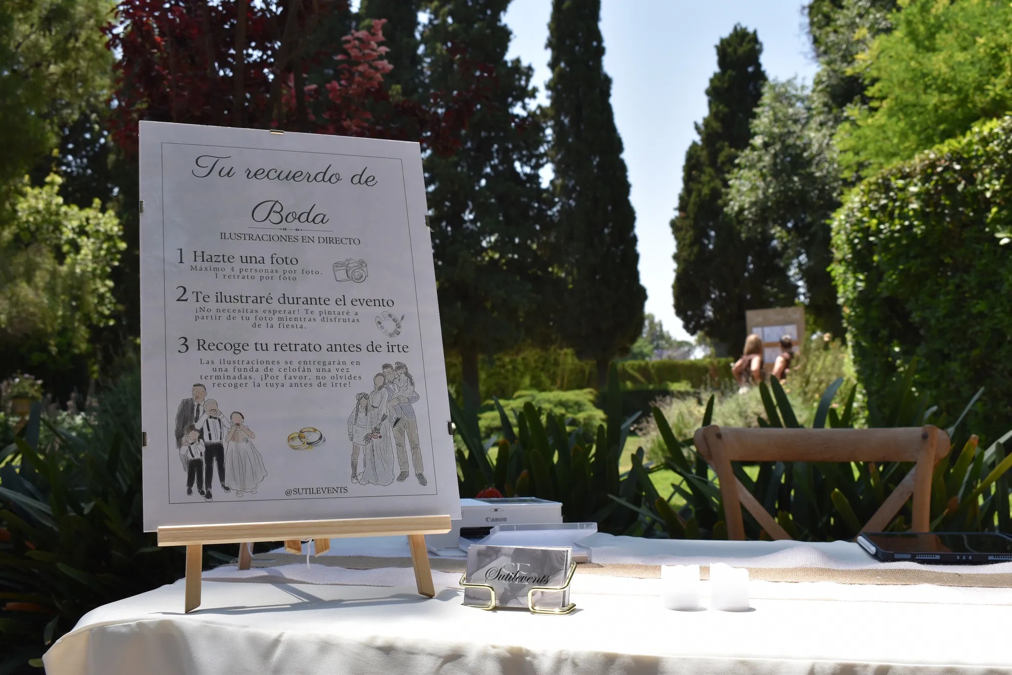 Cartel «Tu recuerdo de boda · ilustraciones en directo» en un caballete sobre la mesa, con tarjetas de SutilEvents y un jardín de cipreses detrás