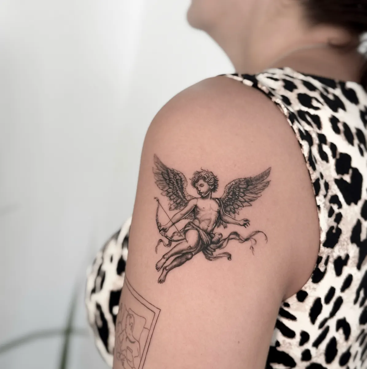 Winged cherub with a bow and flowing ribbon tattooed in black and grey micro-realism on the shoulder